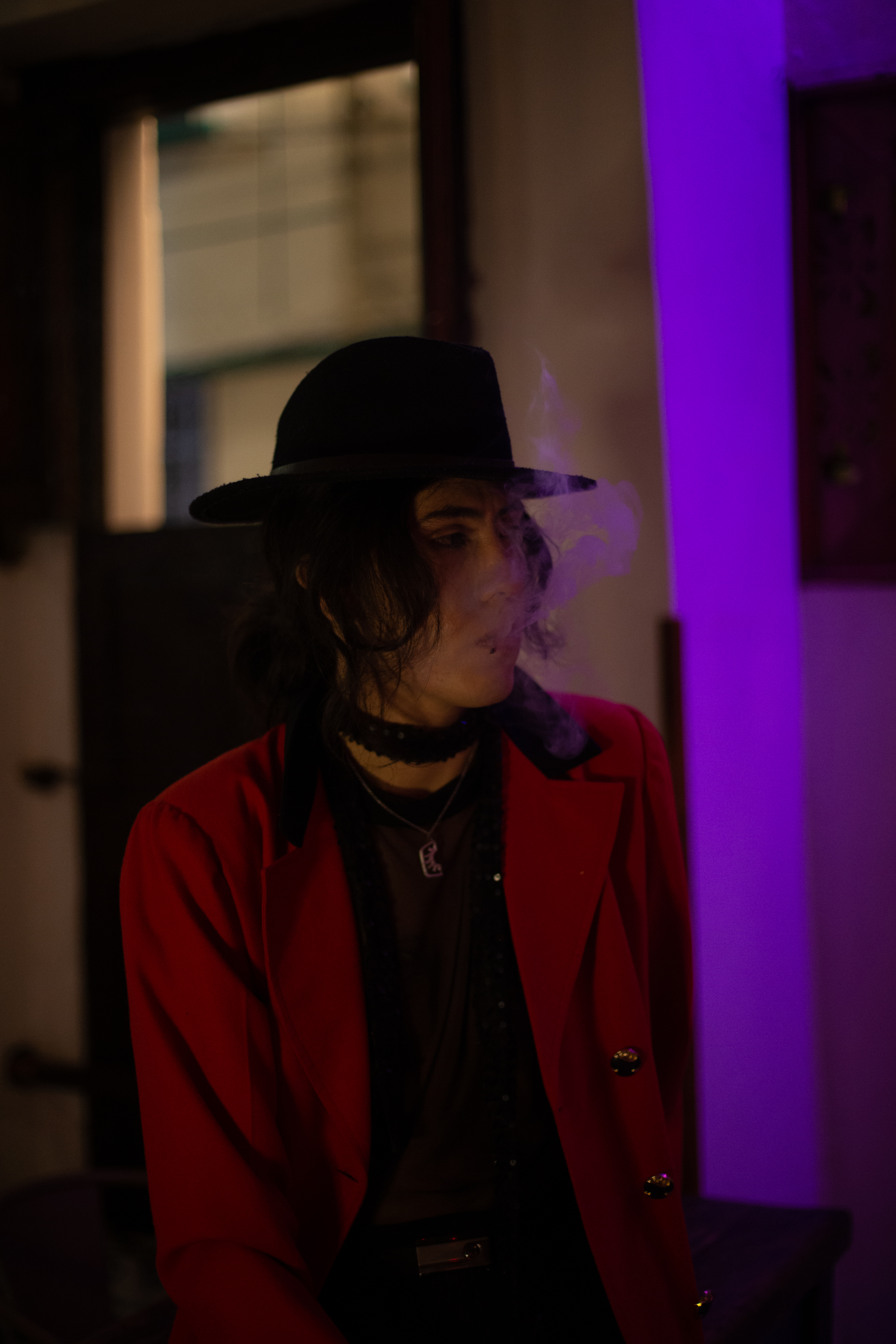 Johan Peraza, atmospheric indoor portrait wearing a wide-brim hat and red blazer in dim purple light